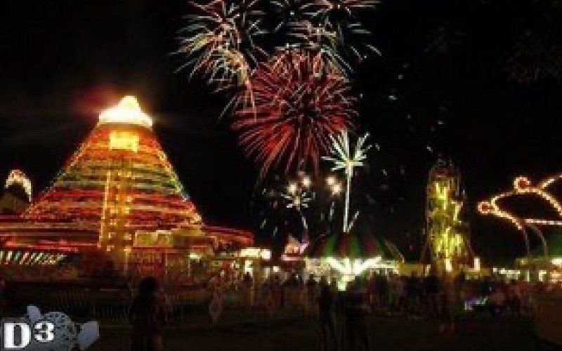 Fireworks bursting over a lit pyramid-shaped ride and Ferris wheel at an amusement park at night