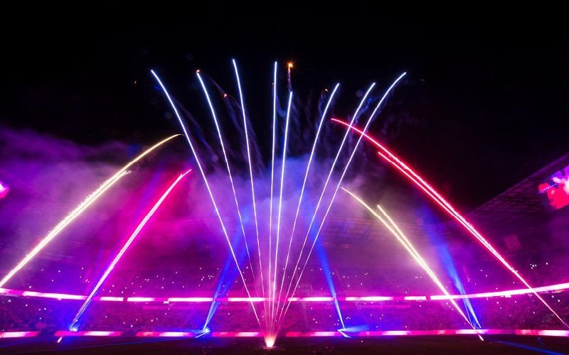 Pink, blue and gold comet streaks fanning up from a stadium field, with haze and colored light along the stands