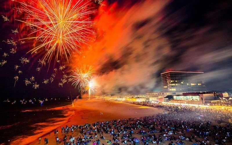 Orange and gold fireworks over the Asbury Park beach, with a large crowd on the sand and a hotel at right