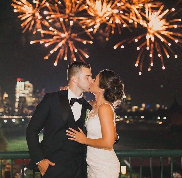 Couple kissing on a balcony at night as orange fireworks burst over a lit city skyline behind them