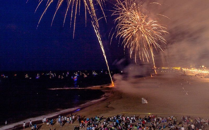 Gold fireworks and a rising comet over a beach at dusk, with a crowd seated on the sand and boats offshore
