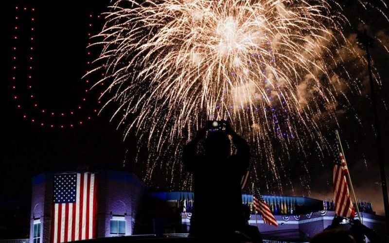 Silhouetted spectator photographing large gold fireworks above a building draped with a flag, with drone lights at left