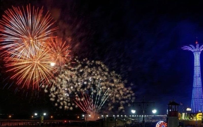 Orange and gold fireworks over a boardwalk beside a lit blue tower at an amusement area at night