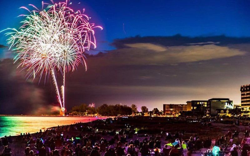 Pink, white and green firework burst over a crowded beach at dusk, with a glowing waterline
