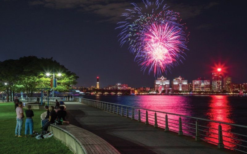Pink and blue fireworks over a river and city skyline, with people on a lawn and a waterfront walkway
