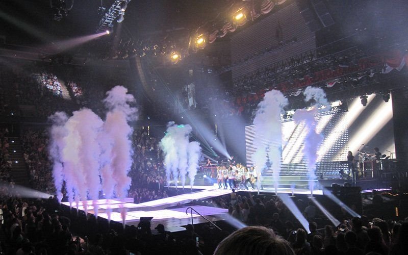Columns of white vapor rising along a concert stage runway, lit by beams of light, with a crowd in the arena