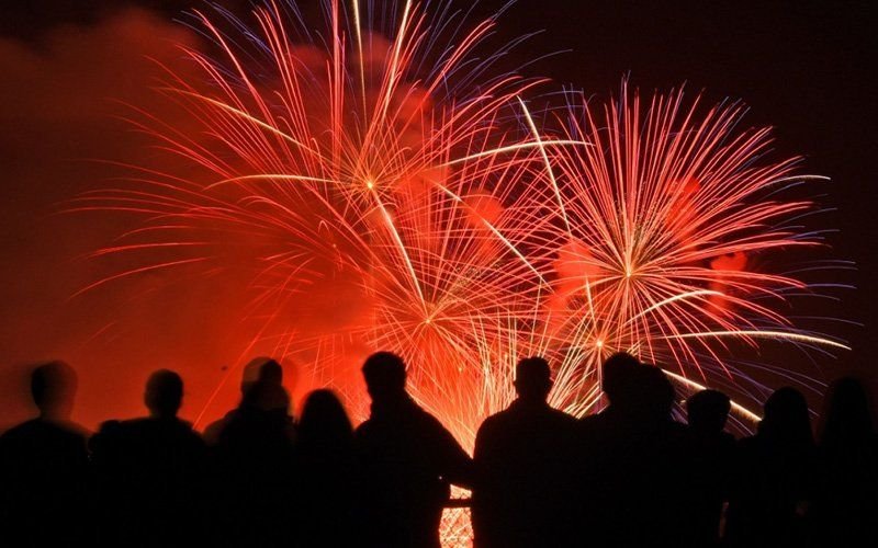Silhouetted crowd watching red and orange fireworks burst against a glowing night sky