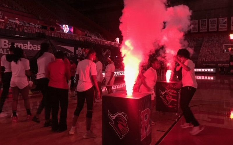 Pink-lit vapor and a flame jet beside a cabinet on an arena floor as people gather during a pre-game show