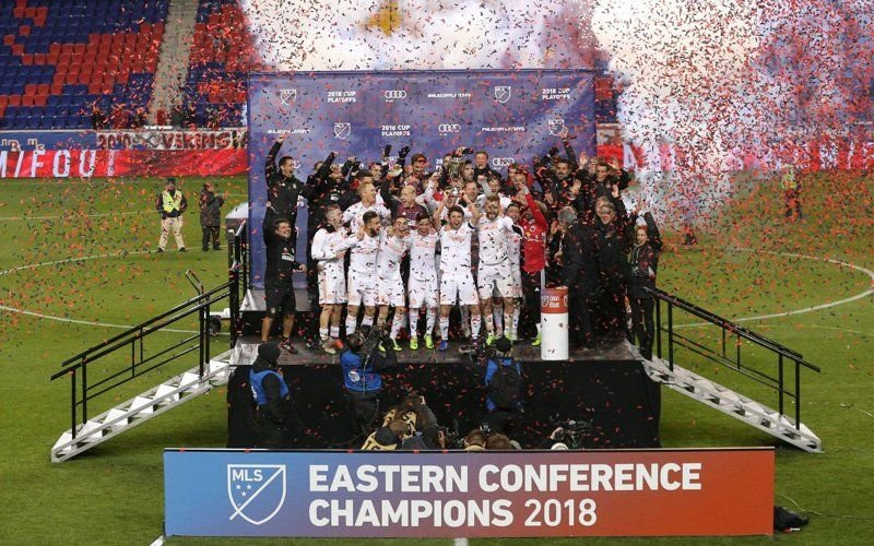 Soccer players on a podium at midfield as red confetti fills the air, under a banner for a conference title