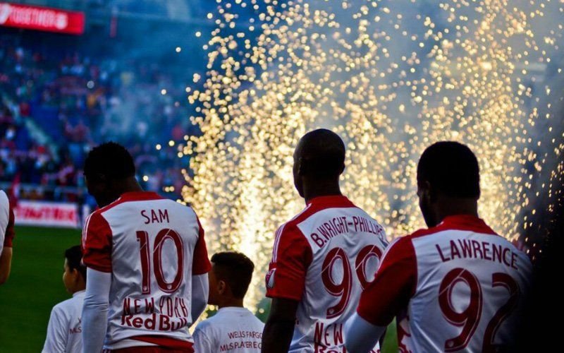 Players in white jerseys facing a curtain of gold gerb sparks at a stadium player entrance