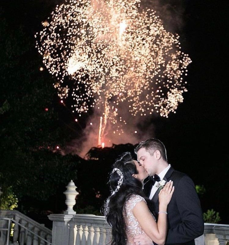 Bride and groom kissing beside a stone balustrade at night under a large gold firework burst