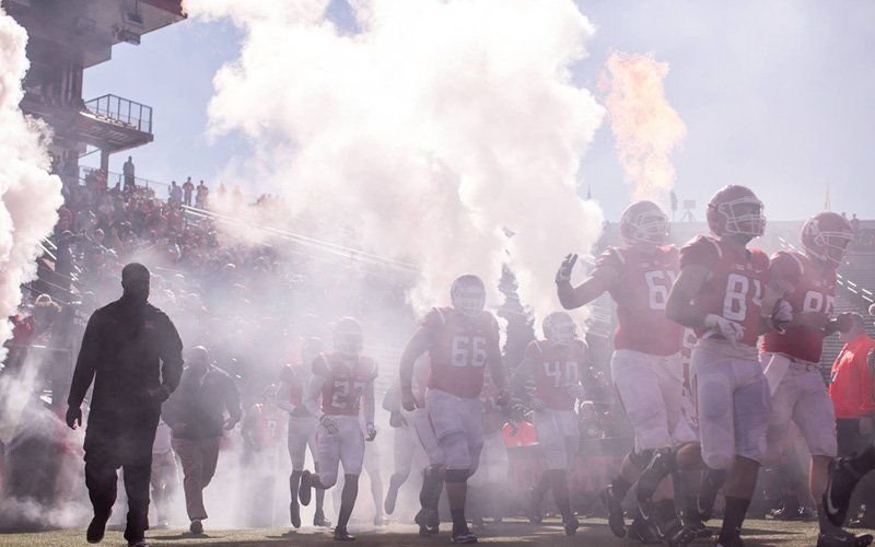 Football players in red jerseys running out through white smoke as a flame burst rises beside the tunnel