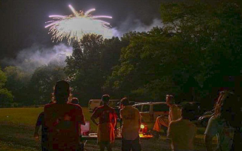 Spectators and parked vehicles on a grass field watching a white firework burst above the trees at night