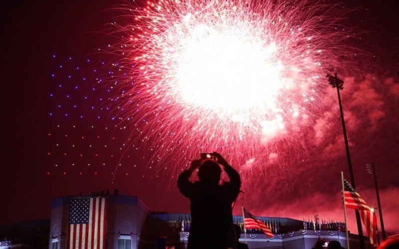 Pink firework burst over a stadium with a US flag, as a spectator in silhouette photographs it