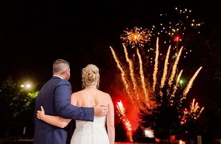 Couple arm in arm watching gold and red fireworks burst over trees at a night wedding
