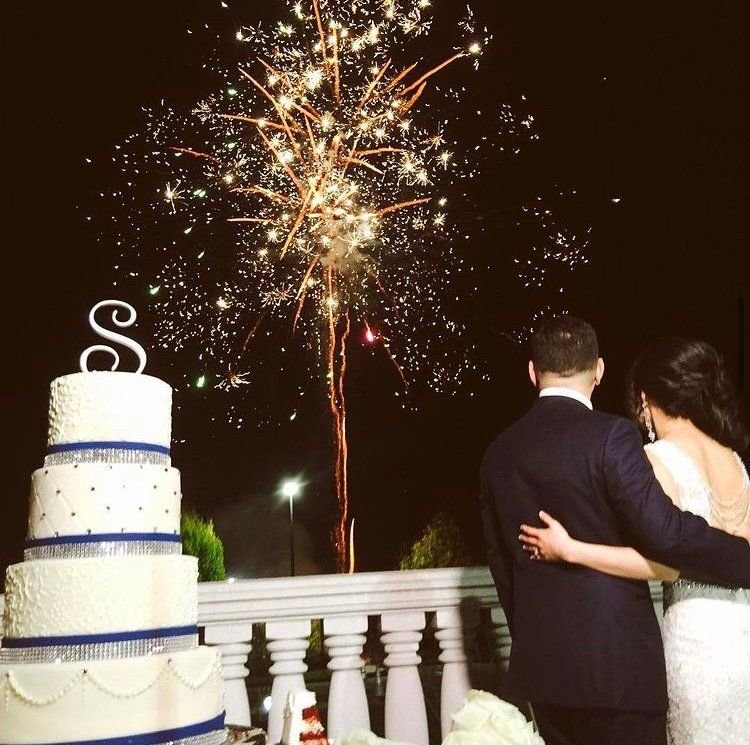 Couple watching a gold firework burst from a terrace at night, with a tiered white wedding cake in the foreground